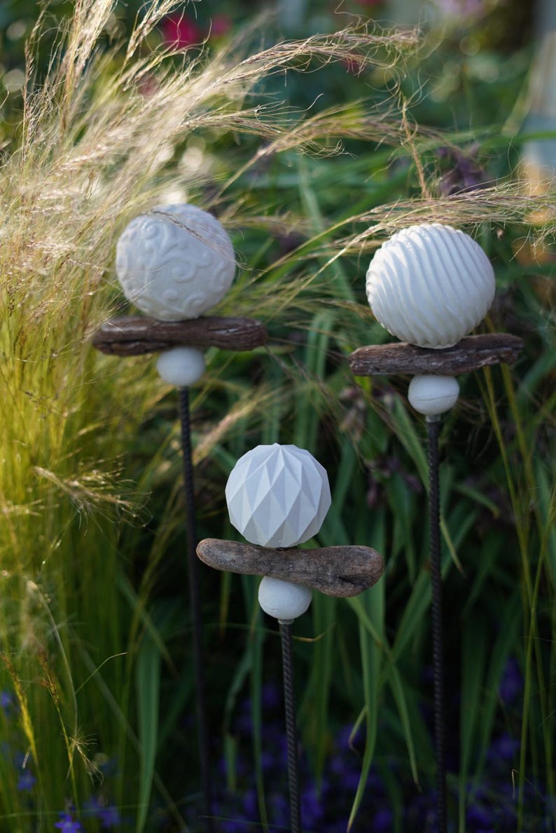 May include: Three garden ornaments with white spherical designs. Each stake has a different textured sphere: one with swirls, one with ridges, and one with a geometric pattern. The spheres are mounted on driftwood and supported by a white ball.