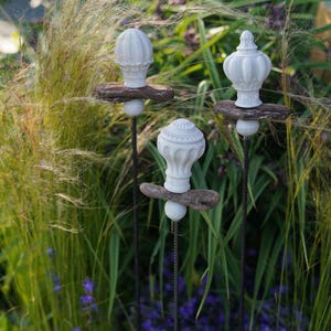 Könnte beinhalten: Drei weiße dekorative Gartenstecker mit verzierten Aufsätzen. Jeder Stecker hat eine weiße, strukturierte Spitze, ein kleines Holzakzent und eine weiße Kugel. Die Stecker sind in einem Garten mit hohem Gras und lila Blumen aufgestellt.