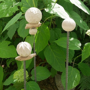 May include: Three garden stakes with white, spherical ornaments. The stakes are made of metal and are set amongst green foliage. The ornaments have different textures and designs, including carved patterns and geometric shapes.