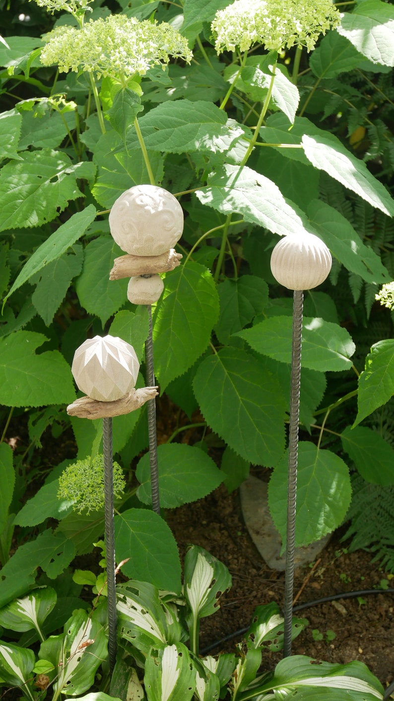 May include: Three garden stakes with white, spherical ornaments. The stakes are made of metal and are set amongst green foliage. The ornaments have different textures and designs, including carved patterns and geometric shapes.