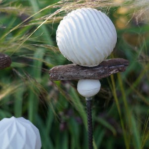 May include: White garden ornaments with a spiral and geometric design. They are mounted on wooden pieces and metal rods, set against green foliage and tall grasses. A unique outdoor decor. The ornaments are a stylish addition to any garden.