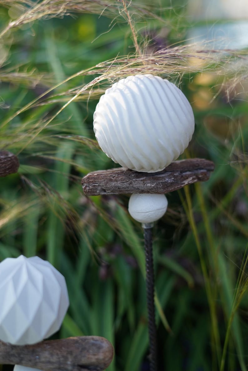 May include: White garden ornaments with a spiral and geometric design. They are mounted on wooden pieces and metal rods, set against green foliage and tall grasses. A unique outdoor decor. The ornaments are a stylish addition to any garden.