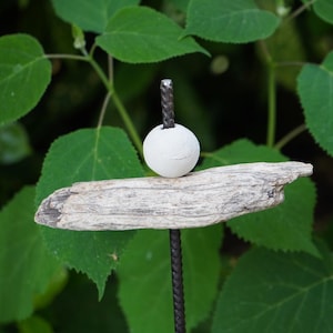May include: A rustic garden stake featuring a white sphere atop a piece of driftwood, mounted on a dark metal rod. The driftwood is a light grey colour, and the background is blurred green foliage.