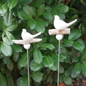 May include: Two white ceramic birds perched on driftwood branches, each attached to a metal stake. The birds are facing different directions and are positioned against a backdrop of green foliage.