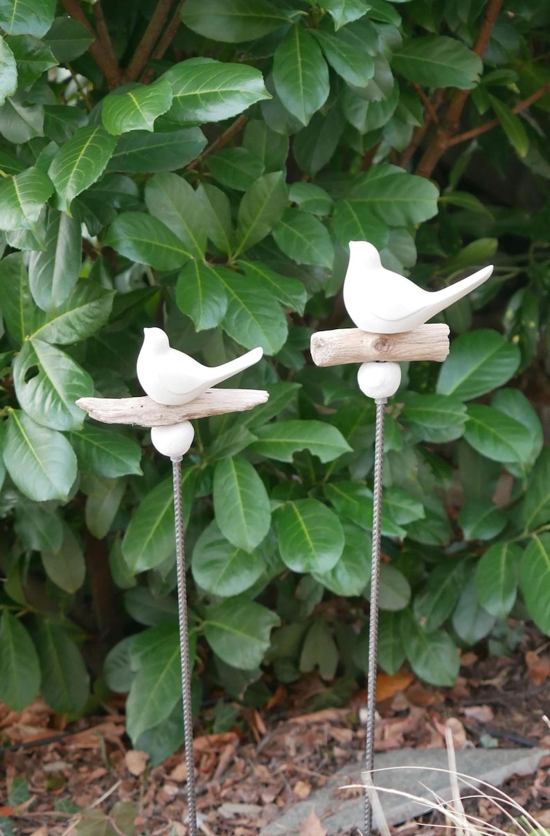 May include: Two white ceramic birds perched on driftwood branches, each attached to a metal stake. The birds are facing different directions and are positioned against a backdrop of green foliage.