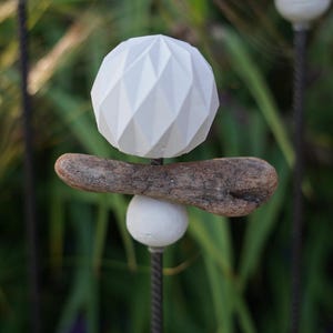 May include: A garden ornament with a white geometric sphere above a piece of driftwood, balanced on a smaller white sphere. The stake is made of twisted metal. The background is a blur of green and purple.