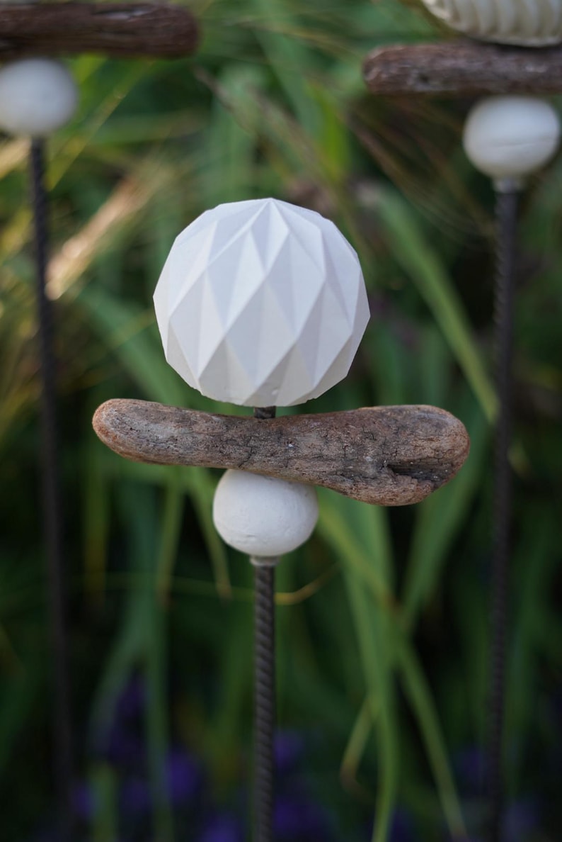 May include: A garden ornament with a white geometric sphere above a piece of driftwood, balanced on a smaller white sphere. The stake is made of twisted metal. The background is a blur of green and purple.