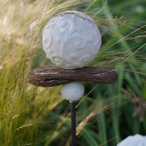 May include: A garden ornament with a white, patterned sphere on a piece of weathered wood. A smaller white sphere is below, attached to a dark metal rod. The background shows tall, green grasses.
