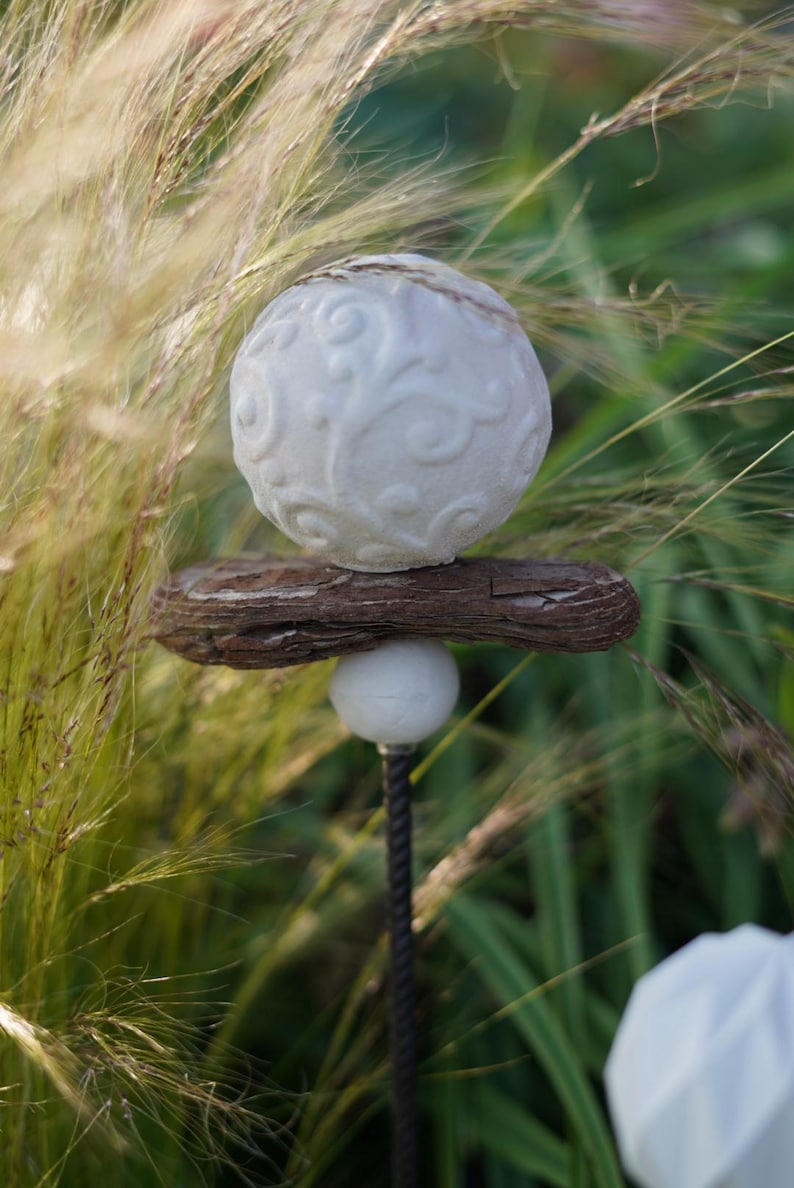 May include: A garden ornament with a white, patterned sphere on a piece of weathered wood. A smaller white sphere is below, attached to a dark metal rod. The background shows tall, green grasses.