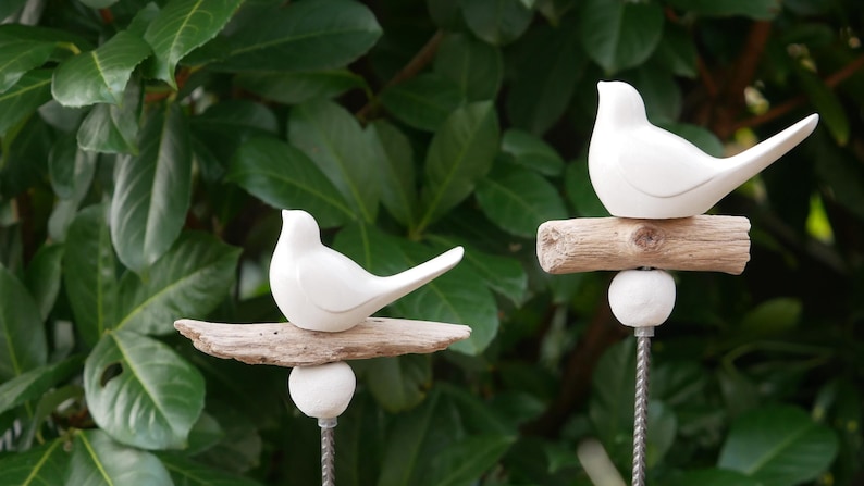 May include: Two white ceramic birds perched on driftwood branches. The branches are attached to metal stakes, creating a decorative garden stake set.