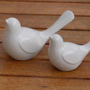 May include: Two white ceramic dove figurines, one with its wings spread out and the other with its wings folded, on a wooden surface.