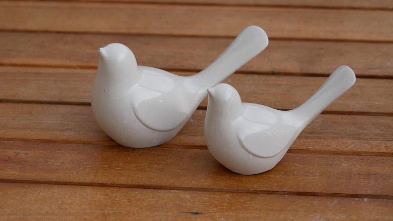 May include: Two white ceramic dove figurines, one with its wings spread out and the other with its wings folded, on a wooden surface.