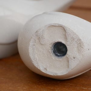 May include: Close-up of the bottom of a white ceramic bird figurine. The bottom has a circular hole with a dark gray plug.