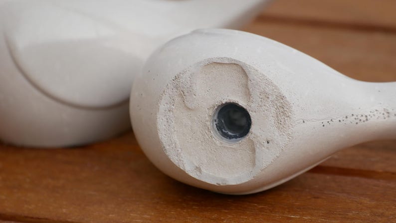 May include: Close-up of the bottom of a white ceramic bird figurine. The bottom has a circular hole with a dark gray plug.