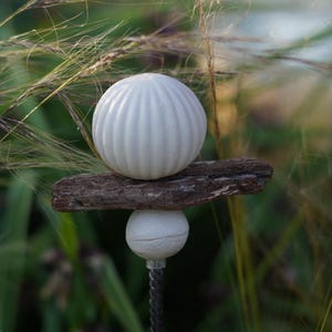 May include: A garden ornament with a white, ribbed sphere balanced on a piece of wood, above a smaller white sphere. The stake is made of twisted metal. The background is a blur of green and brown.