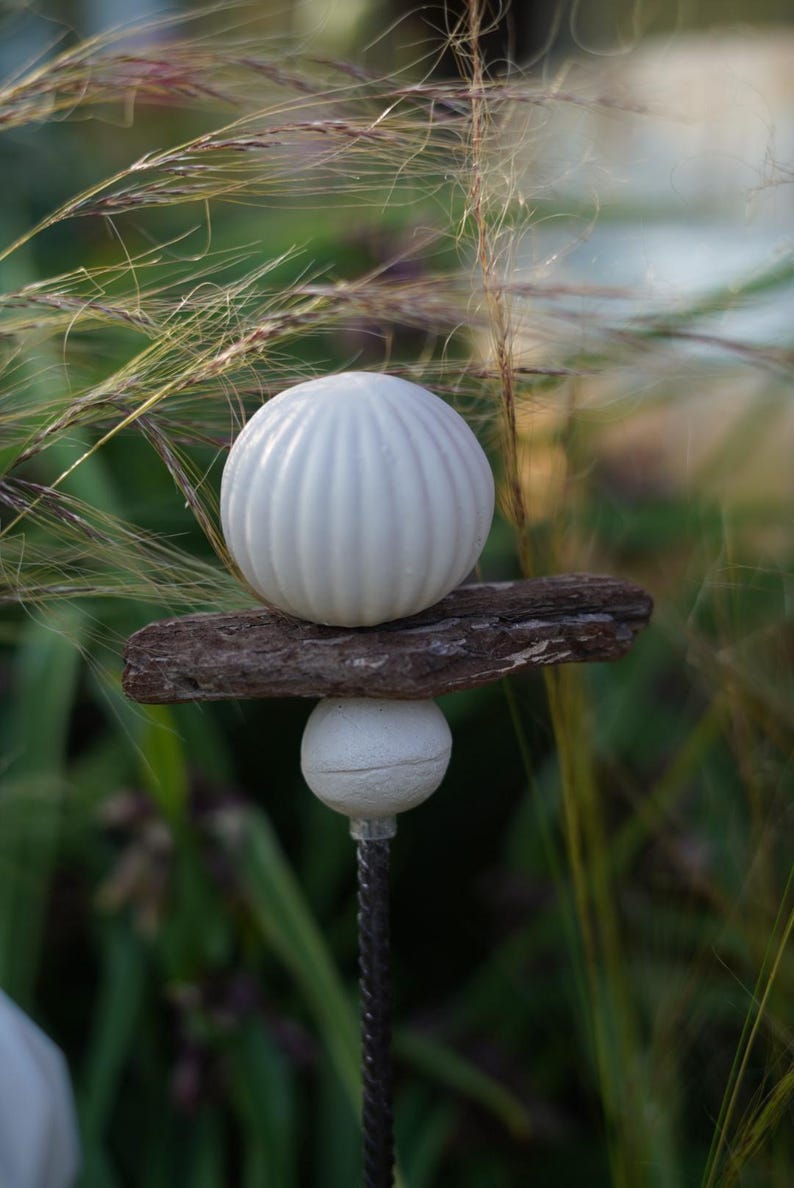 May include: A garden ornament with a white, ribbed sphere balanced on a piece of wood, above a smaller white sphere. The stake is made of twisted metal. The background is a blur of green and brown.