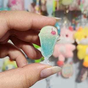 May include: A clear acrylic charm shaped like a soda float. The charm features a gradient of colors, including blue, green, and white, with a cherry on top. The charm is held in a hand, with other charms in the background.
