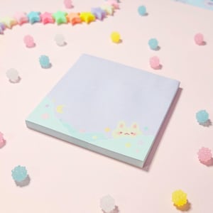 May include: A pastel blue notepad with a cute bunny rabbit illustration and a moon and stars design. The notepad is surrounded by pastel-colored candy sprinkles.