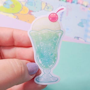 May include: A sticker of a green and blue soda float with a cherry on top. The soda float is in a glass with a clear base and is surrounded by bubbles.