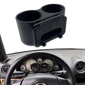 May include: A black dual cup holder accessory featuring a rectangular mounting plate with a horizontal slot, shown above a car dashboard interior that includes a steering wheel and a gauge cluster with two circular AEM brand gauges installed.