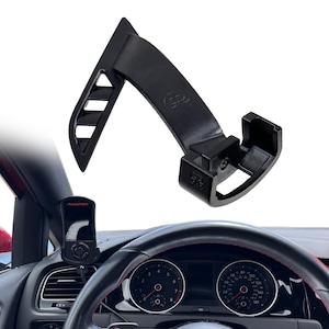 May include: A black plastic car dashboard mount. The mount has a curved design with a slotted section and a rectangular base. The mount is designed to hold a device, such as a performance monitor, near the steering wheel.