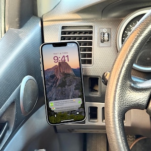 May include: A smartphone displaying a mountain landscape wallpaper is mounted in a car. The screen shows the time as 9:41 AM and a notification. The phone is silver and is mounted on the dashboard near the steering wheel.