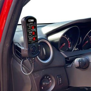 May include: A black Cobb Accessport device mounted in a car's dashboard air vent. The device displays real-time engine data with red and white text on a black screen. The car's dashboard is black and red.