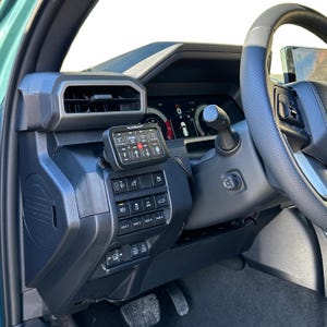 May include: Interior view of a vehicle's dashboard, featuring a black control panel with multiple buttons and switches. A small digital display with the text "AUXBEAM" is mounted on the dashboard. The steering wheel and other interior components are also visible.