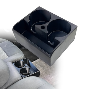May include: A black plastic car cup holder insert featuring two circular beverage compartments and a rectangular storage slot. The unit is designed with a central divider and a small circular opening between the two main cup holders.