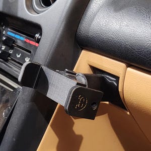 May include: A black 3D-printed car phone mount installed in a vehicle's dashboard. The mount is attached to the dashboard and glove compartment. The interior of the car has black and tan elements.
