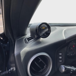 May include: A black circular AEM OBD-II gauge mounted in a housing on the textured black dashboard of a vehicle, positioned near a circular air vent and a set of control buttons labeled km/h MPH, ODO TRIP, and DISP on the instrument panel.