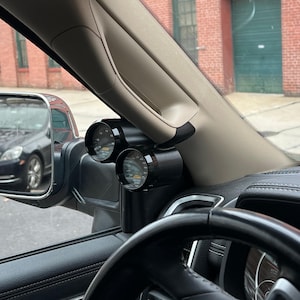 May include: Black automotive gauges mounted on a vehicle's interior pillar. The gauges display readings with black faces, white markings, and orange needles. The gauges are labeled "FUEL" and "WATER TEMP". The steering wheel and side mirror are also visible.