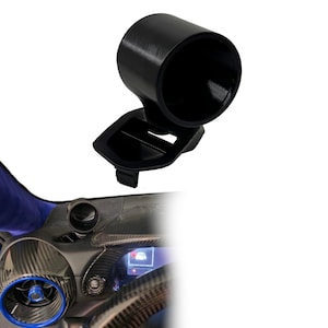 May include: A black cylindrical gauge pod mount designed for automotive dashboard installation. The mount features a circular housing for a gauge and a flat base with a clip mechanism for secure attachment to a vehicle's air vent or dashboard surface.