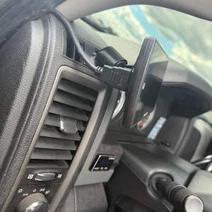May include: A black electronic device is mounted on a car's dashboard, connected by a black cable. The device is positioned near the air vent and steering wheel. The car's interior features a black and gray color scheme.