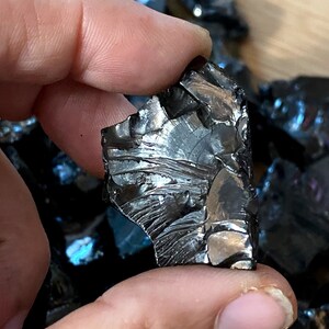 May include: A piece of black obsidian with iridescent blue and green hues. The obsidian is rough and has a jagged edge. It is a natural stone and has a unique, irregular shape.