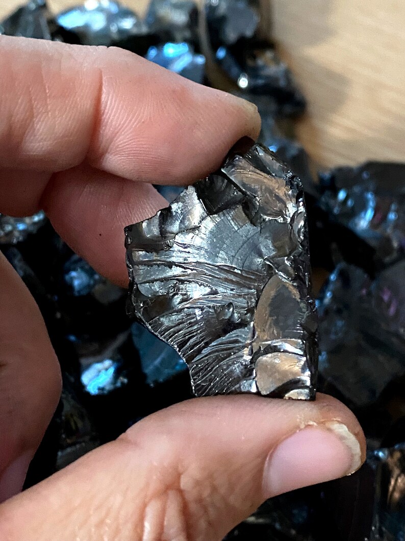 May include: A piece of black obsidian with iridescent blue and green hues. The obsidian is rough and has a jagged edge. It is a natural stone and has a unique, irregular shape.
