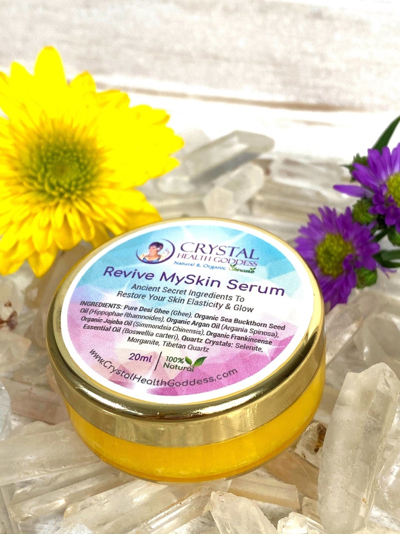 May include: A small yellow jar with a gold lid labeled "Revive MySkin Serum" with a list of ingredients including pure desi ghee, organic sea buckthorn seed oil, organic argan oil, organic jojoba oil, and organic frankincense essential oil. The jar is sitting on a bed of clear quartz crystals.