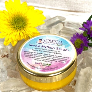 May include: A small, gold-rimmed jar of yellow serum labeled "Revive MySkin Serum" with the text "Ancient Secret Ingredients To Restore Your Skin Elasticity & Glow" and a list of ingredients. The jar is sitting on a bed of clear quartz crystals.