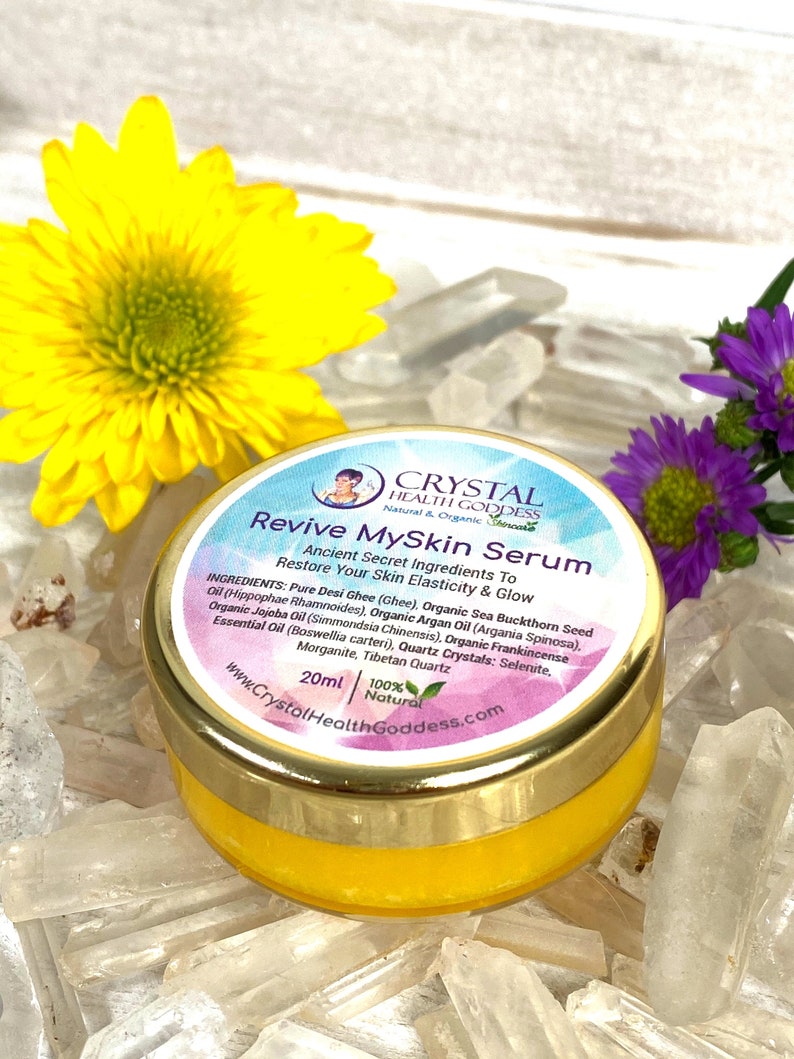 May include: A small, gold-rimmed jar of yellow serum labeled "Revive MySkin Serum" with the text "Ancient Secret Ingredients To Restore Your Skin Elasticity & Glow" and a list of ingredients. The jar is sitting on a bed of clear quartz crystals.