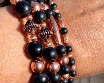 May include: A set of four bracelets featuring black and copper beads. The bracelets are stacked on a person's wrist. The copper beads are shiny and have a metallic finish. The black beads are matte and have a smooth surface. The bracelets are a mix of round and cylindrical beads.