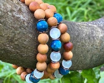May include: A bracelet made with wooden beads, blue and white gemstone beads. The bracelet is on a tree branch with green foliage in the background. @CrystalHealthGoddess