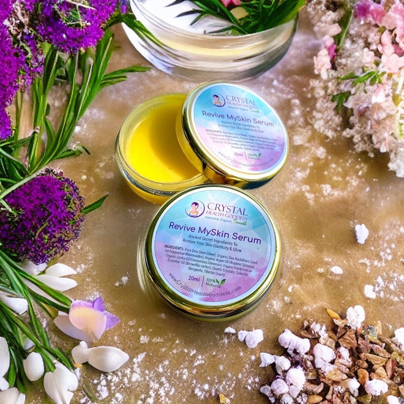 May include: Two small jars of yellow serum with gold lids. The jars are labeled "Revive MySkin Serum" and feature a logo with a woman's face and the text "Crystal Health Goddess". The jars are sitting on a light brown surface with white powder and small white flowers.