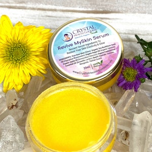 May include: A small, round jar with a gold lid and a yellow label. The label reads "Crystal Health Goddess Natural & Organic" and "Revive MySkin Serum". The jar contains a yellow serum and is sitting on a white surface with clear crystals.