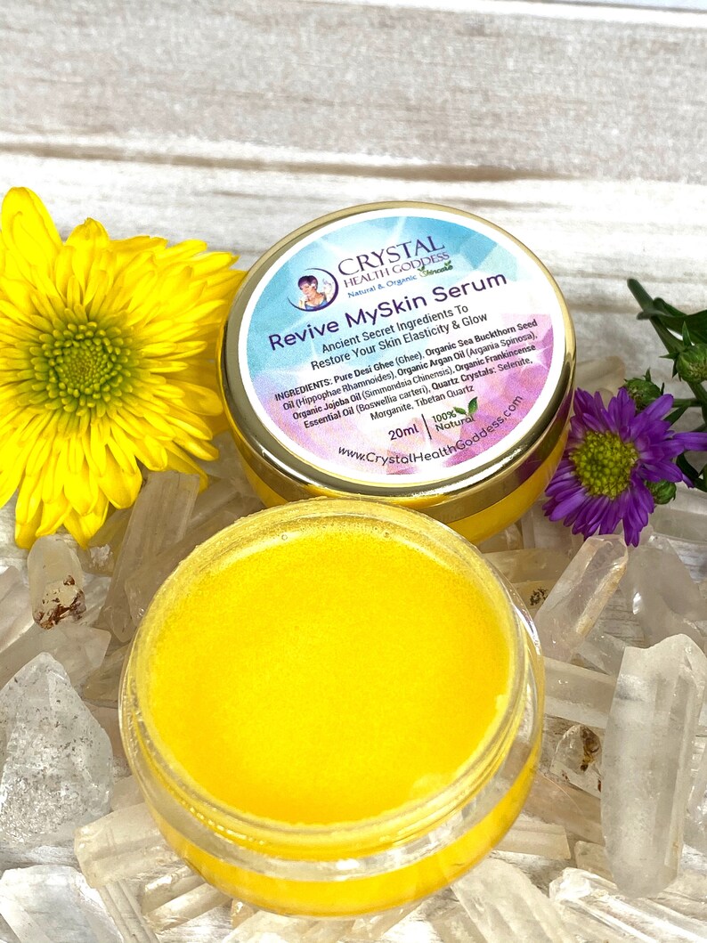 May include: A small, round jar with a gold lid and a yellow label. The label reads "Crystal Health Goddess Natural & Organic" and "Revive MySkin Serum". The jar contains a yellow serum and is sitting on a white surface with clear crystals.