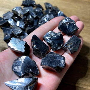 May include: A collection of black obsidian stones with a reflective, iridescent surface. The stones are various sizes and shapes, with some having a rough, jagged texture.