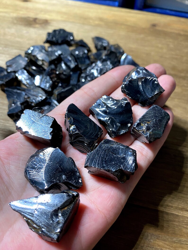 May include: A collection of black obsidian stones with a reflective, iridescent surface. The stones are various sizes and shapes, with some having a rough, jagged texture.