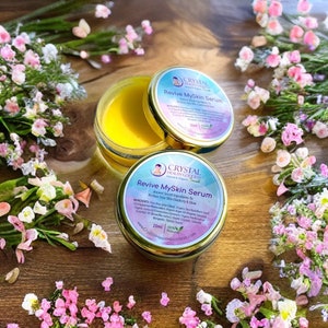May include: A small, gold-colored jar with a yellow cream inside. The jar is labeled "Revive MySkin Serum" and has a logo with a woman's face and the words "Crystal Health Goddess". The jar is surrounded by pink and white flowers on a wooden surface.
