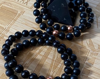 May include: Two black beaded bracelets with copper accents on a wooden surface. The bracelets are stacked on top of each other. The word "RELAX" is engraved on the wooden surface.