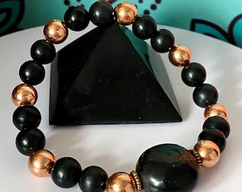 May include: Black and copper beaded bracelet with a large black bead in the center. The bracelet is resting on a black pyramid.