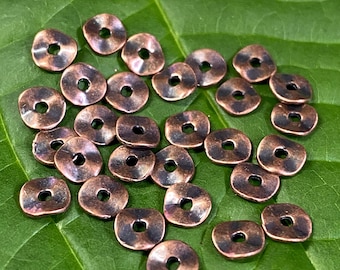 May include: A collection of copper-toned metal beads with a slightly rounded, flat shape. The beads have a hole in the center and are scattered on a green leaf.
